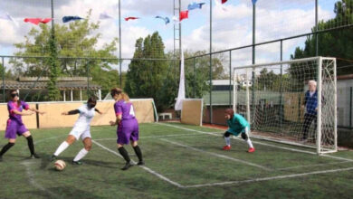 Bir futbol sahasında, görme engelli kadın futbolcular gözlerine siyah bir bant takılı şekilde futbol oynuyorlar. Sahada 4 sporcu olduğu görülüyor. Beyaz formalı kadın futbolcu topu ayağıyla kaleye doğru sürüyor. Önünde ve arkasında mor renkli forma yiyen rakip takım futbolcuları savunma yapmaya çalışıyor.