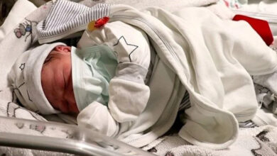 Fotoğrafta, bir bebek küvöz içinde yatıyor. Bebeğin yüzü kısmen kapatılmış ve üzerine ince bir örtü örtülmüş.