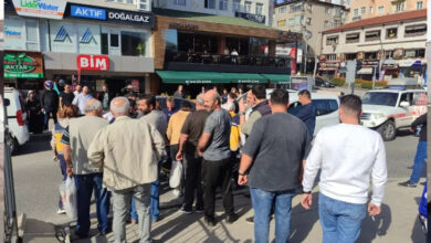 Bir cadde üzerinde araçların durduğu ve kalabalık bir grup insan bir alanda toplanmış. Toplanan kalabalığın arasında ambulans görevlileri ve polisin olduğu görülmektedir.