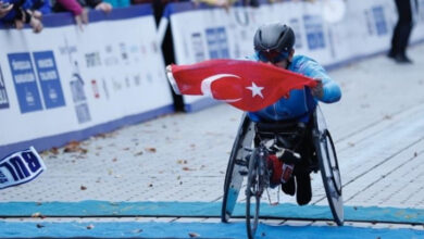 Fotoğrafta, önde bir ve yanlarda iki tekeri olan özel yapan tekerlekli sandalye ile engelli birey maraton koşusunu bitirirken Türk bayrağını çıkartarak bitiş çizgisini geçiyor.