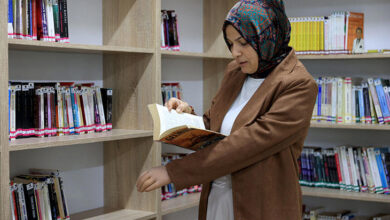 Fotoğrafta, kütüphanede kitap okuyan bir öğretmen var. Öğretmen ayakta kitabı incelerken protez olan sol kolunu kitabın altında tutarak yukarıda kalması için destek veriyor.