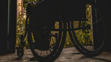 Fotoğrafta, tekerlekli sandalye kullanan engelli birey karanlık bir odadan kapıyı açarak yeşil ve gün ışığını olduğu bir bahçeye doğru çıkıyor.