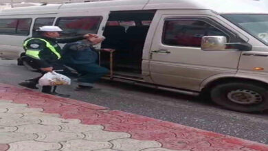 Minibüsün önünde binmek için hareket eden görme engelli birey ve arkasında polis var. Polis memuru görme engelli bireyin kafasını eliyle kapatarak yağmurdan ıslanmasını engelliyor.