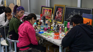 Fotoğrafta engelli bireyler tabure üzerinde oturarak önlerindeki masada yer alan resim malzemeleri ile resim yapıyorlar. Masanın üzerinde tamamlanmış resimlerde yan yana dizilmiş.
