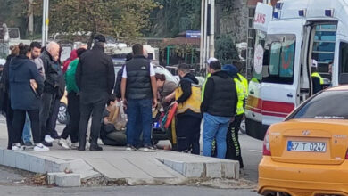 Fotoğrafta, yol kenarında kaldırımda oturan bir kişinin etrafına kalabalık bir insan grubu toplanmış. Kalabalık arasında sağlık personelleri var. Yolun kenarına park etmiş bir ambulans aracı var.