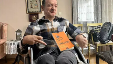 Tekerlekli sandalye kullanıcısı Gökhan Arslan, yazmış olduğu kitabı kucağında duruyor ve kameraya gülümseyerek bakıyor.