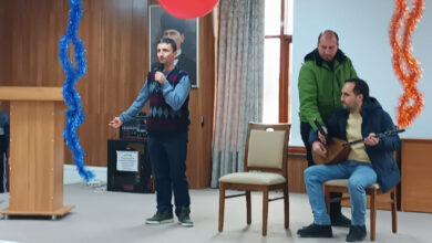 Fotoğrafta, bir sahnede yer alan engelli bireyler var. Engelli bireylerden birinin elinde mikrofon var ve şarkı söylüyor. Diğer engelli birey ise tahta bir sandalyede oturuyor ve elinde tuttuğu bağlamayı çalıyor.