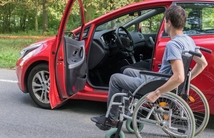 Tekerlekli sandalye kullanan bedensel engelli bir adam kırmızı renkli otomobillerinin sürücü koltuğu kapısına doğru yaklaşıyor ve sürücü koltuğu kapısı açık.