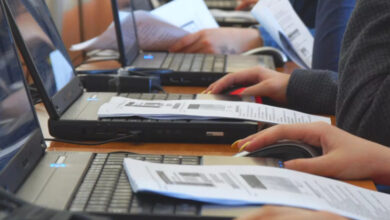 Fotoğrafta, bir masanın üzerinde yan yana dizilmiş dizüstü bilgisayarlar var. Bilgisayar başında ise birkaç kişinin elleri gözüküyor.