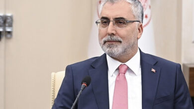 Çalışma ve Sosyal Güvenlik Bakanı Vedat Işıkhan, bir toplantıda önünde mikrofonlar yer alıyor ve çevresindeki konuşmaları dinliyor.