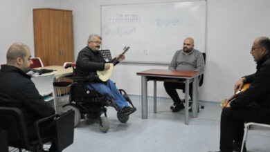 Görselde, bir sınıfta bulunan dört yetişkin erkek yer alıyor. Sol tarafta bir kişi, tekerlekli sandalyesinde otururken bağlama çalıyor. Diğer üç kişi, sandalye veya masada oturarak ona odaklanmış durumda. Arka planda bir beyaz tahta görülüyor, üzerinde notalar ve müzikle ilgili çizimler var. Ortam sade bir sınıf atmosferinde, duvarlar beyaz ve dekor minimal. Bu görüntü, bir müzik eğitimi veya ders ortamını yansıtıyor.