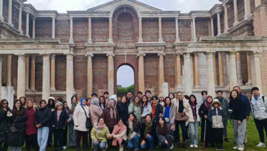 Fotoğrafta, büyük bir grup insan tarihi bir yapının önünde poz veriyor. Arka planda, sütunları ve kemerli girişleriyle dikkat çeken, antik bir mimariye sahip büyük bir yapı yer alıyor. İnsanlar kışlık kıyafetler giymiş, bazıları atkı ve bereler takmış. Grupta farklı yaş gruplarından bireyler bulunuyor; bazıları gülümseyerek poz verirken, bazıları daha ciddi bir ifadeye sahip. Görme engelli bireyler beyaz baston kullanıyor. Fotoğraf, tarihi ve kültürel bir gezi etkinliği sırasında çekilmiş gibi görünüyor.