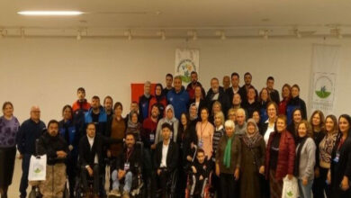 Fotoğrafta, bir grup insan geniş bir salonda bir araya gelmiş şekilde görülüyor. Çeşitli yaş gruplarından bireyler, hem engelli bireyler hem de onların destekçileri olarak, birlikte poz vermişler. Grupta tekerlekli sandalyedeki kişiler de dikkat çekiyor ve toplumun her kesiminden bireylerin bir araya geldiği bir etkinlik ortamı olduğu anlaşılıyor. Arka planda Osmangazi Kent Konseyi ve Engelliler Meclisi'nin logoları yer alırken, Türk bayrağı da karede görünmekte. Etkinliğin amacı, "Afetlerde Engelsiz Birlik" gibi bir başlıkla engellilerin afet durumlarında yaşadığı zorluklara yönelik farkındalık yaratmak ve çözüm üretmek olarak öne çıkıyor. Toplumsal dayanışma ve birlik mesajı veren anlamlı bir anı yansıtıyor.