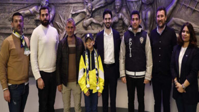 Fotoğrafta, bir grup insan bir arada poz veriyor. Arka planda kabartma bir sanat eseri bulunuyor; eser spor temalı figürlerden oluşmuş gibi görünüyor. Grubun ortasında, sarı-lacivert Fenerbahçe renklerinde forma ve atkı giyen bir çocuk dikkat çekiyor. Çocuğun etrafında çeşitli kişiler yer almakta: sivil giyimli bireyler, bir polis yeleği giyen kişi ve takım elbise giymiş başka bir kişi var. Fotoğraf samimi bir etkinlik anını yansıtıyor; Fenerbahçe taraftarı olan bu çocuğa bir jest yapılmış.