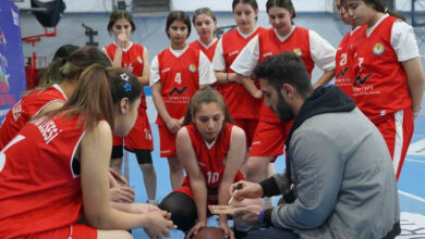 Fotoğrafta, kapalı bir spor salonunda duvar kenarına yakın bir alanda dizin üzerine çökmüş erkek antrenör ve çevresinde yaşları 13-15 arasında değişen kız sporcular var. Antrenör elindeki kağıtla yazılı taktikleri sporcularına anlatıyor.