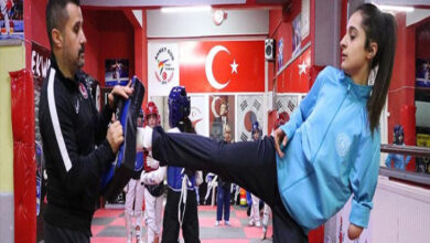 Bir spor salonunda çekilen bu görselde, 17 yaşındaki engelli sporcu Tuana Çelik, tekvando antrenmanında güçlü bir tekme hareketi sergiliyor. Sağ kolu ampute ve azimli bir duruş sergileyen sporcu, mavi eşofman üstü giymiş. Sol tarafta antrenör, elinde tekvando hedef yastığı tutarak dikkatle sporcusunu yönlendiriyor. Arka planda Türk bayrağı, tekvando ile ilgili posterler ve antrenman yapan diğer sporcular yer alıyor. Görsel, Tuana Çelik’in spordaki azmi ve başarı hedefini yansıtıyor.