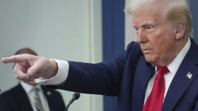 ABD Başkanı Donald Trump, koyu lacivert bir takım elbise ve kırmızı kravat giymiş, sağ eliyle işaret ederek kararlı bir ifadeyle bir yere bakıyor. Arka planda bulanık bir kişi görülüyor.