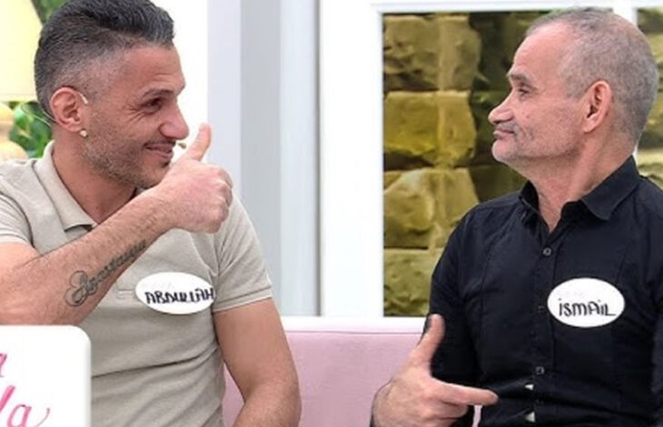 Görselde, bir televizyon programında çekilmiş bir sahne yer almakta. İki erkek, pembe bir koltukta oturuyor ve birbirlerine el işaretleriyle iletişim kuruyor. Soldaki kişi açık renk bir tişört giymiş ve üzerinde "Ramazan" yazılı bir isim etiketi bulunuyor. Sağdaki kişi ise siyah bir gömlek giymiş ve etiketi "İsmail" olarak görünüyor.