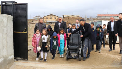 Fotoğrafta, bir grup ilkokul öğrencisi ve yetişkinler açık bir kapıdan içeri giriyor. Çocuklar kışlık montlar giymiş, bazıları neşeyle gülümsüyor. Ön tarafta bir adam, siyah akülü bir tekerlekli sandalyeyi tutuyor. Arkalarında takım elbiseli yetkililer ve köy evleri görülüyor. Bulutlu bir hava hakim.