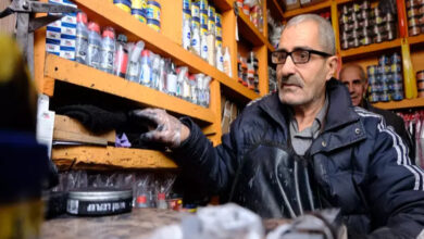 Görselde, gözlüklü ve orta yaşlı bir adam, bir ayakkabı boyacısı tezgâhında çalışırken görülüyor. Üzerinde lacivert bir mont ve önlük bulunuyor, elleri eldivenli ve boyalı. Arka planda, turuncu raflarda dizilmiş çeşitli boya ve bakım malzemeleri yer alıyor. Adam, tezgâhın arkasında bir şeye uzanırken odaklanmış bir ifadeye sahip. Arkasında başka bir kişi daha var, ancak net bir şekilde görünmüyor.