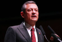 Fotoğrafta, CHP Genel Başkanı Özgür Özel, siyah takım elbise ve kırmızı desenli bir kravat giymiş şekilde mikrofona konuşuyor. Koyu bir arka plan önünde duruyor ve resmi bir etkinlikte konuşma yapıyor gibi görünüyor.