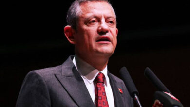 Fotoğrafta, CHP Genel Başkanı Özgür Özel, siyah takım elbise ve kırmızı desenli bir kravat giymiş şekilde mikrofona konuşuyor. Koyu bir arka plan önünde duruyor ve resmi bir etkinlikte konuşma yapıyor gibi görünüyor.