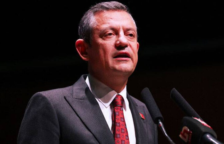 Fotoğrafta, CHP Genel Başkanı Özgür Özel, siyah takım elbise ve kırmızı desenli bir kravat giymiş şekilde mikrofona konuşuyor. Koyu bir arka plan önünde duruyor ve resmi bir etkinlikte konuşma yapıyor gibi görünüyor.