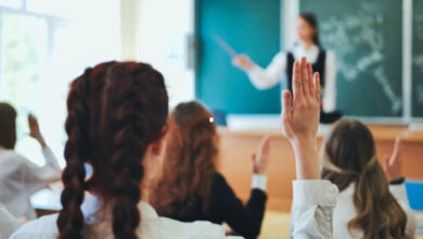 Bu görselde bir sınıf ortamı görülüyor. Ön planda, sıralarda oturan birkaç öğrenci ellerini kaldırmış, öğretmenden söz almak istiyorlar. Öğrenciler genellikle okul forması giymiş. Arka planda ise öğretmen, kara tahta önünde ders anlatıyor ve elindeki bir işaret çubuğuyla tahtadaki yazıları gösteriyor. Sınıf oldukça aydınlık, pencere kenarından gelen doğal ışık ortamı ferahlatıyor.