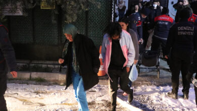 Görsel, gece saatlerinde bir yangın tahliyesini gösteriyor. Ön planda yaşlı bir kadın başörtüsü ve siyah mont giymiş halde yürürken yanında genç bir kadın bulunuyor. Arkalarında itfaiye ekipleri ve kalabalık bir grup insan var. Zeminde beyaz bir madde (muhtemelen yangın söndürme köpüğü) görülüyor. Görselde "Denizli İtfaiyesi" yazılı üniformalı kişiler de bulunuyor.