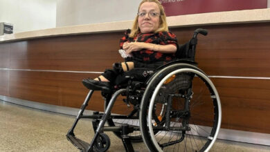 Fotoğrafta, tekerlekli sandalyede oturan bir kadın görülüyor. Sarı saçlı ve gözlüklü olan kadın, kırmızı-siyah desenli kısa kollu bir elbise giymiş. Sol kolu protez olup sağ elinde bir kağıt parçası tutuyor. Arka planda "Ahmed Adnan Saygun Sanat Merkezi" yazılı bir tabela bulunan modern bir resepsiyon alanı yer alıyor.