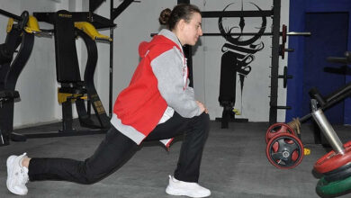 Görselde, spor salonunda bir kadın ısınma hareketi yapıyor. Üzerinde kırmızı ve gri renkli bir spor ceketi, siyah eşofman altı ve beyaz spor ayakkabıları var. Kadın, bir bacağını öne atarak esneme hareketi gerçekleştiriyor. Arka planda ağırlık kaldırma ekipmanları ve duvarda siyah bir judo figürü resmi bulunuyor.