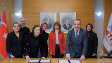 Fotoğrafta, resmi bir ortamda bir grup insan bir araya gelmiş. Ön planda iki kişi, bir masa üzerinde bulunan belgeleri imzalarken gülümsüyor. Arkalarında dört kişi daha bulunuyor. Masanın arkasında Türk bayrağı, Hitit Üniversitesi logosu bulunan bir bayrak ve duvarda iki siyah-beyaz portre yer alıyor.