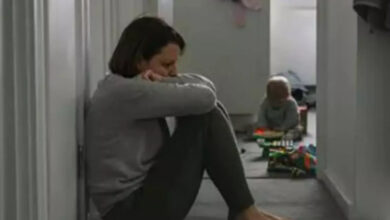 Görselde, bir kadın koridorda duvara yaslanarak yere oturmuş, üzgün bir şekilde başını kollarına yaslamış duruyor. Uzakta, başka bir odada bir bebek oyuncaklarla oynuyor. Ortam loş ve hüzünlü bir atmosfer hâkim. Kadının yüzündeki ifade, duygusal bir sıkıntıyı veya çaresizliği yansıtıyor.