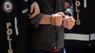 Görselde, polis ekipleri tarafından kelepçelenmiş bir şüpheli görülüyor. Şüphelinin elleri kelepçeli şekilde öne doğru tutulmuş ve yanında resmi polis yeleği giyen bir görevli bulunuyor. Görüntü, bir gözaltı anını yansıtıyor.