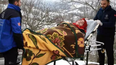 Görselde, karlı bir kırsal bölgede iki sağlık görevlisi, sedyede yatan yaşlı bir kadını hastaneye taşımak için hazırlıyor. Kadın, başörtüsü takıyor ve üzerine kalın desenli bir battaniye örtülmüş. Görevliler, Kocaeli Başiskele Belediyesi’nin "Mavi Ambulans" hizmeti kapsamında hastayı güvenli bir şekilde taşımaya çalışıyor. Arka planda karla kaplı ağaçlar ve doğal bir manzara görülüyor.