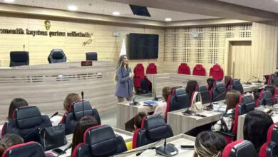 Fotoğrafta, modern bir konferans veya toplantı salonunda gerçekleştirilen bir eğitim görülüyor. Salon, ahşap kaplamalar ve kırmızı-siyah renklerde modern koltuklarla döşenmiş. Katılımcılar, mikrofonlu masalarda oturmuş, eğitimi dinliyorlar. Eğitimi veren kişi, salonun ortasında ayakta duruyor ve gri bir ceket giymiş. Salonun ön kısmında, belediye meclis salonlarına özgü bir kürsü ve arkasında üç büyük koltuk bulunuyor. Duvara ise "Egemenlik, kayıtsız şartsız milletindir." yazısı asılmış, yanında Türkiye Cumhuriyeti'nin amblemi yer almakta. Arka planda bir Türk bayrağı dikkat çekiyor. Bu görüntü, işitme engelli bireylerin hizmetlerden daha iyi yararlanabilmesi için belediye personeline verilen işaret dili eğitimine ait. Katılımcılar dikkatle dinlerken, ortam oldukça resmi ve eğitim odaklı görünüyor.