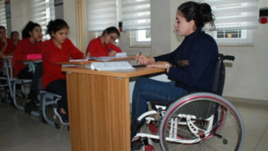 Görselde, tekerlekli sandalyede oturan bir kadın, sınıfta bir sınava veya derse katılıyor. Masasında kitaplar ve notlar bulunuyor. Arka planda, kırmızı üniformalı diğer öğrenciler de sıralarında oturmuş, yazı yazıyor. Sınıf, geniş pencereleri ve modern düzeniyle aydınlık bir ortam sunuyor.