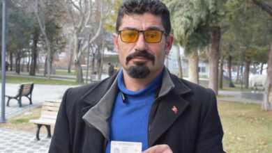 Görselde, bir adam açık havada bir parkta duruyor ve elinde kimlik kartını tutuyor. Üzerinde mavi bir kazak ve siyah bir kaban var. Sarı tonlu gözlükler takıyor ve bıyıklı, sakallı bir görünüme sahip.