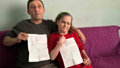 Görselde, bir adam ve bir kadın mor renkli bir koltukta oturuyor. Adam koyu renk bir kazak giymiş ve elinde bir belge tutuyor. Kadın ise kırmızı bir kıyafet giymiş, saçlarını yeşil bir toka ile toplamış ve gözleri kapalı bir şekilde belgesini gösteriyor. Adam, kadına destek olmak amacıyla kolunu omzuna koymuş.