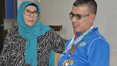 Görselde, başörtülü ve gözlüklü bir kadın ile spor kıyafetleri giymiş bir erkek yan yana oturuyor. Erkek, boynunda çeşitli madalyalar taşımakta ve gülümsüyor. Kadın, ona destekleyici bir ifadeyle bakıyor ve kolunu omzuna atmış durumda. Arka planda sade bir ev ortamı görülüyor.