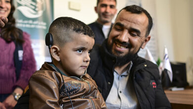 Fotoğrafta, işitme cihazı takılı küçük bir çocuk ve ona gülümseyerek bakan bir adam görülüyor. Çocuk kahverengi bir ceket giyerken, adam siyah mont ve açık renkli bir gömlek giymiş. Arkalarında birkaç kişi daha bulunuyor ve ortam resmi bir kurum ya da sağlık merkezi gibi görünüyor. Çocuğun ciddi bir ifadesi varken, yanındaki adam mutlu ve gururlu bir şekilde gülümsüyor.
