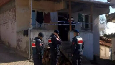 Görselde, tek katlı eski bir evin önünde görev yapan jandarma ekipleri görülüyor. Üç jandarma personeli siyah bere ve üniformalarıyla dikkat çekerken, biri telsiziyle iletişim kuruyor. Evin giriş kapısına mavi-beyaz şerit çekilmiş, içeride ise birkaç görevli daha bulunuyor. Ev eski ve yıpranmış görünüyor, duvarlarında çatlaklar var. Merdivenlerin altında odun yığınları yer alıyor.