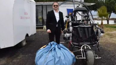 Görselde, şık bir takım elbise ve güneş gözlüğü takan bir adam, motorlu bir yamaç paraşütü (paramotor) ve yanında büyük bir paraşüt çantasıyla poz veriyor. Arka planda küçük bir yapı, ağaçlar ve açık hava ortamı görülüyor. Zeminin toprak ve çakıl karışımı olduğu anlaşılıyor.