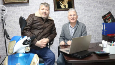 Fotoğrafta, ofis ortamında iki adam görülüyor. Biri deri ceketli ve mavi pantolonlu olarak motosikletin yanında oturuyor, diğeri ise gri ceket ve gözlükleriyle masada, dizüstü bilgisayarın başında bulunuyor. Arka planda taş desenli duvar, Atatürk portresi ve kitaplarla dolu bir raf yer alıyor. Masada bir hesap makinesi ve ofis malzemeleri de mevcut.