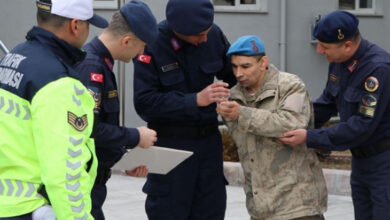 Fotoğrafta, jandarma ve trafik polisleri üniformalı bir grup görevli, asker kıyafeti giymiş bir engelli bireye yardımcı oluyor. Engelli birey mavi bereli ve kamuflajlı, yanında duran jandarmalar ona destek vererek eline bir belge veya madalya tutuşturuyor. Görevlilerden biri elinde bir dosya tutuyor. Arka planda gri bir bina görülüyor. Fotoğraf, engelli bireyin askerlik hayalinin gerçekleştiği özel bir anı yansıtıyor.