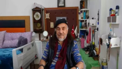 Görselde, yaşlı bir adam masa başında oturuyor ve kameraya gülümsüyor. Uzun saçlı ve sakallı olan adam, siyah bir bere, mavi tonlarında bir gömlek ve bordo bir atkı takıyor. Arka planda, bir bakım merkezi odasını andıran bir ortam bulunuyor. Sol tarafta hastane tipi bir yatak, duvarda büyük bir saat ve küçük raflarda çeşitli eşyalar yer alıyor.