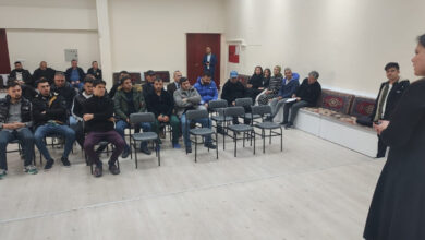 Fotoğrafta bir eğitim veya bilgilendirme toplantısı düzenleniyor. Bir kadın siyah kıyafetiyle ön tarafta konuşma yaparken, karşısında sandalyelere oturmuş bir grup insan dikkatle dinliyor. Katılımcıların bir kısmı sandalyelerde, bazıları ise arka tarafta sedir tarzı oturma alanında oturuyor. Oda geniş ve aydınlık, duvarlar sade, zeminde açık renk parke bulunuyor.