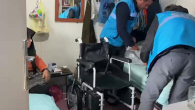 Görselde, mavi yelekli görevliler bir odada engelli bir birey için yardım sağlıyor. Odanın içinde bir tekerlekli sandalye ve yatak görülüyor. Yaşlı bir kadın yatakta otururken, görevliler bazı eşyaları düzenliyor veya yardım sağlıyor gibi görünüyor.