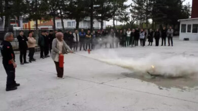 Görselde, açık bir alanda yangın söndürme tatbikatı gerçekleştiriliyor. Başörtülü bir kadın, elinde yangın söndürücü ile küçük bir ateşi söndürmeye çalışıyor. Yanında bir görevli veya eğitmen duruyor. Çevrede, tatbikatı izleyen ve eğitim alan bir grup insan bulunuyor. Arka planda ağaçlar, binalar ve bir güvenlik kulübesi görülebiliyor. Etkinlik, yangınla mücadele eğitimi kapsamında düzenlenmiş gibi görünüyor.