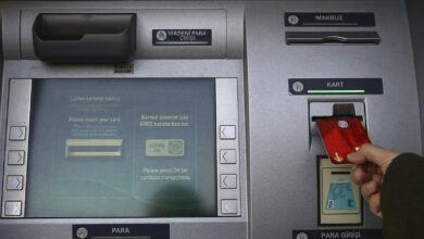 Görselde, bir kişi kırmızı renkte bir banka kartını ATM'ye yerleştiriyor. ATM ekranında kart takılması gerektiğine dair bir uyarı metni yer alıyor. Cihazın üzerinde "Para Girişi," "Makbuz," "Kart" ve "Madeni Para Çıkışı" gibi bölümler bulunuyor.